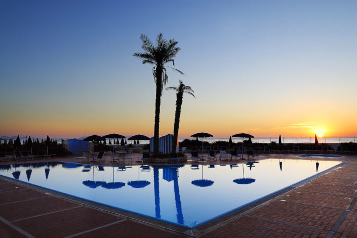 Immagine della piscina di un Hotel a Trapani con le palme, il mare ed il tramonto