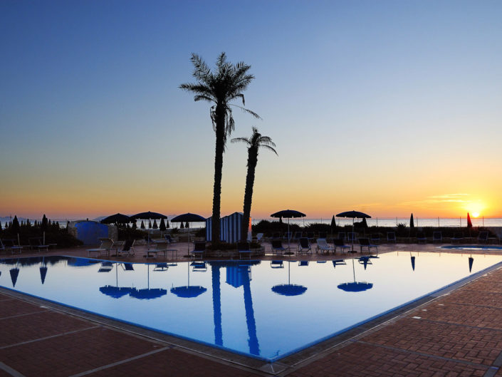 Immagine della piscina di un Hotel a Trapani con le palme, il mare ed il tramonto